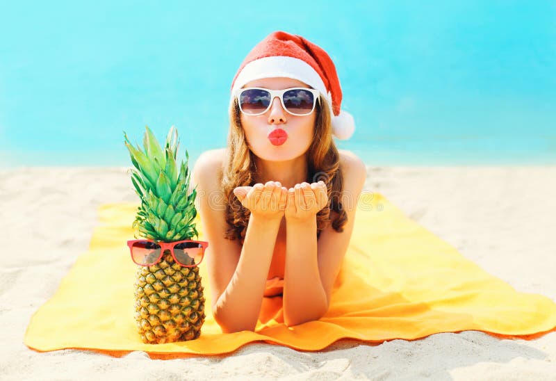 Christmas portrait pretty young woman in red santa hat with pineapple sends air kiss lying on beach over blue sea royalty free stock photo